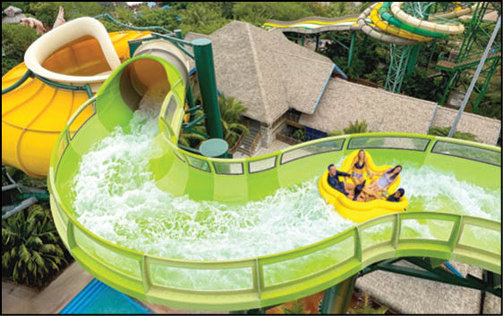 Four people wearing yellow life jackets are riding a yellow raft down a winding green water slide at a water park. Description generated by AI