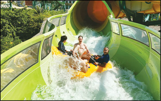 Three adults are riding a yellow inflatable raft down a green enclosed water slide with splashing water around them. Description generated by AI