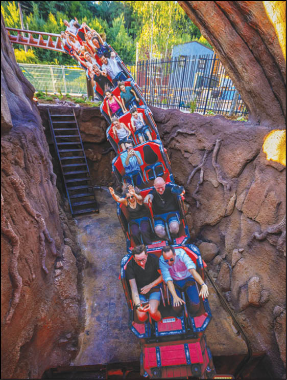 A group of people wearing casual clothing are riding a red roller coaster descending through a rocky tunnel with trees and fencing visible in the background. Description generated by AI