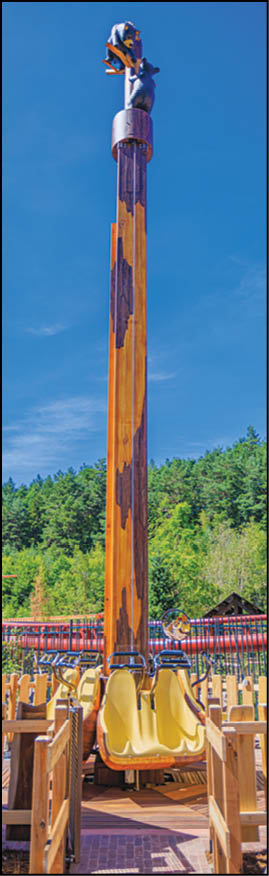 A person wearing a blue outfit is climbing the top of a tall wooden amusement park ride with a yellow slide at the bottom against a clear blue sky. Description generated by AI