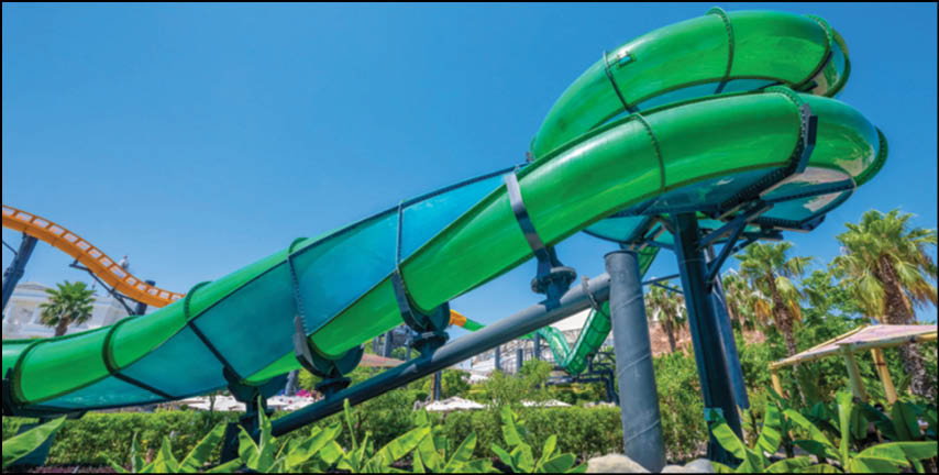 A person is sliding down a large green enclosed water slide surrounded by lush greenery and palm trees under a clear blue sky. Description generated by AI