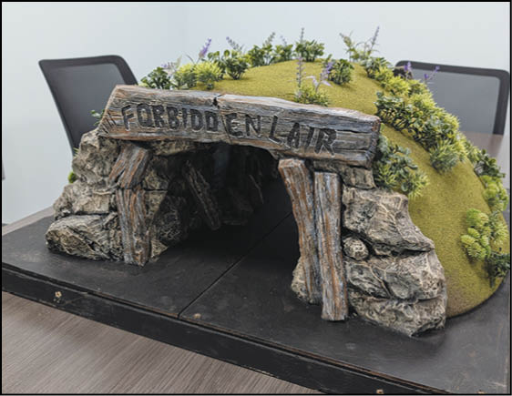 A detailed miniature model of a cave entrance labeled “FORBIDDEN LAIR" with rocky sides and a grassy, flower-covered hill on top, placed on a table with chairs in the background. Description generated by AI