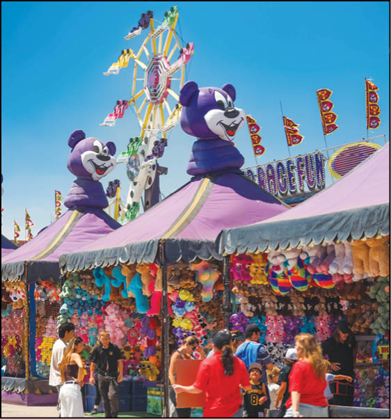 People are gathered around a colorful carnival game booth decorated with large purple bear heads and stuffed animal prizes, with a Ferris wheel and flags visible in the background under a clear blue sky. Description generated by AI