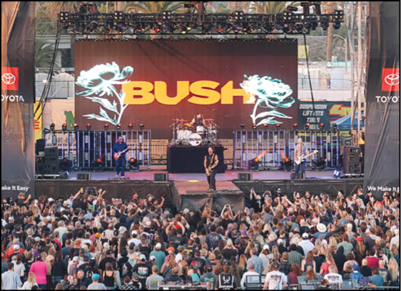 A band performs on an outdoor stage with a large screen behind them displaying the word “BUSH" and floral graphics while a large crowd watches. Description generated by AI