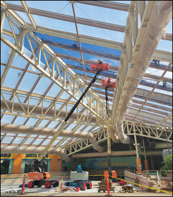 Two people wearing safety gear are working on a large glass ceiling structure while standing on an elevated platform. Description generated by AI