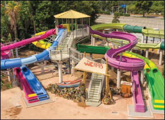 A colorful water park slide structure with multiple twisting and straight slides in pink, blue, green, and yellow surrounding a central platform with stairs and a sign that reads “The Crab." Description generated by AI