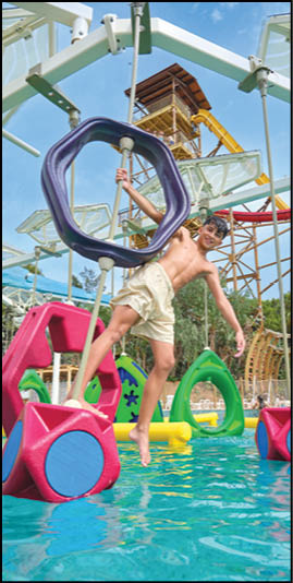 A child wearing shorts is swinging on a large purple ring above a pool with colorful floating play structures and a water park tower in the background. Description generated by AI