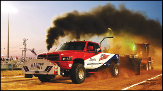 A red and white truck labeled “Bandit" is pulling a heavy sled on a dirt track, emitting thick black smoke from its exhaust. Description generated by AI