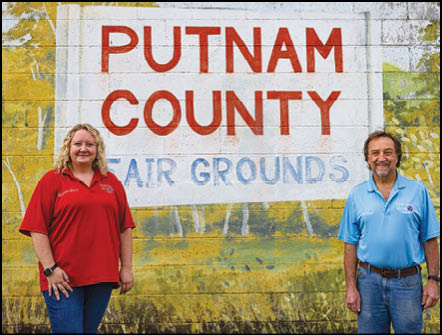 Two adults wearing casual shirts stand smiling in front of a painted wall sign that reads “Putnam County Fair Grounds." Description generated by AI