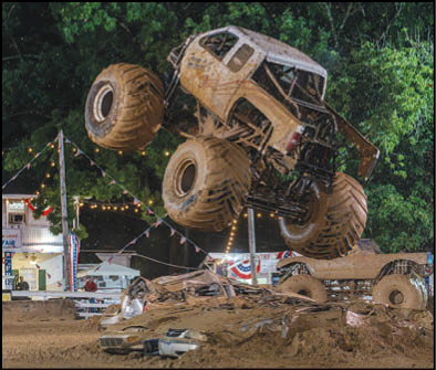 A large monster truck is airborne above a dirt track with crushed cars beneath it and festive lights and trees in the background. Description generated by AI