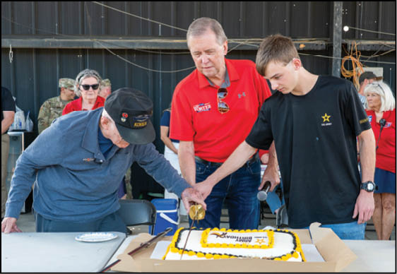 Three people stand around a large rectangular cake with yellow and white frosting, with one person pouring a liquid onto the cake while the others watch. Description generated by AI