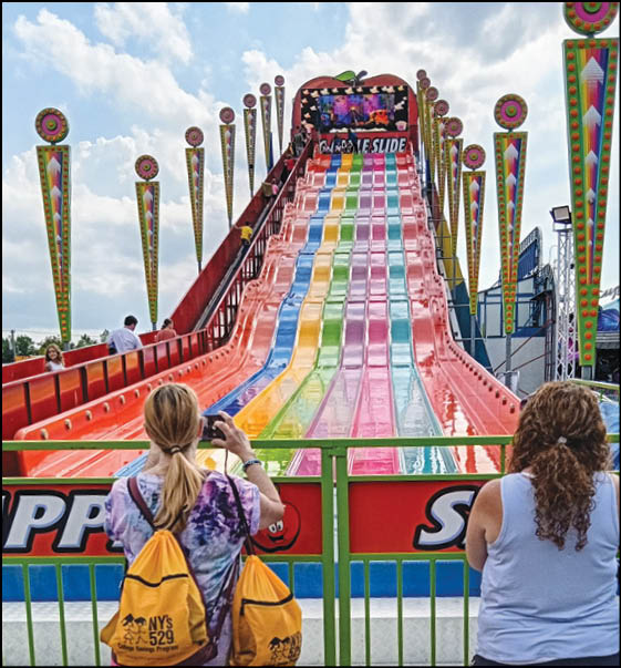 Two people stand at the bottom of a colorful multi-lane slide at an outdoor amusement park or fair. Description generated by AI