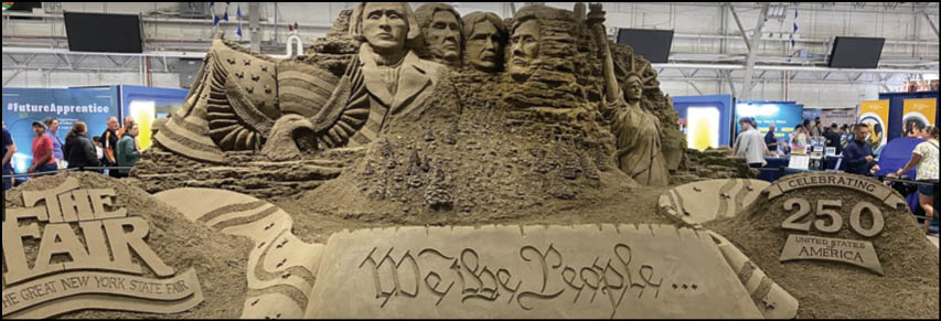 A detailed sand sculpture at a fair features carved faces, an eagle, the phrase “We the People," and signs reading "The Fair" and "250 Years of America" with people and booths visible in the background. Description generated by AI