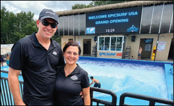 Two adults wearing black polo shirts and sunglasses stand smiling in front of a wave pool with a digital sign announcing the grand opening of EpicSurf USA. Description generated by AI