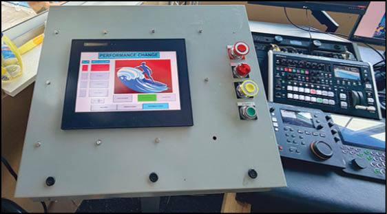 A control panel with a touchscreen displaying a performance change interface and several colored buttons is positioned next to a desk with electronic equipment and monitors. Description generated by AI