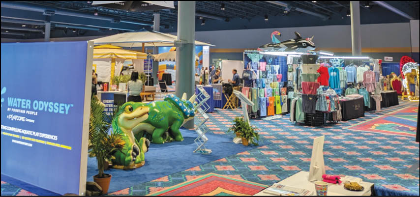 An indoor event space features a colorful carpet, a large green frog sculpture, a booth with clothing displays, and several people walking and sitting. Description generated by AI