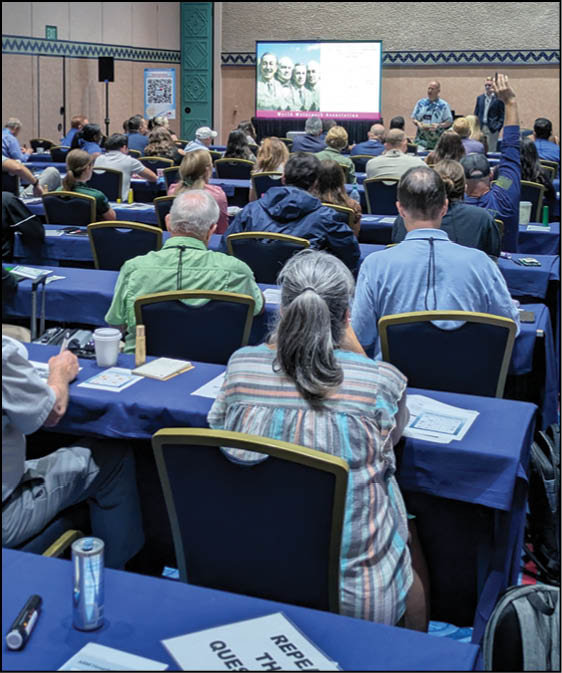 People seated at tables in a conference room are listening to a speaker presenting information displayed on a screen at the front. Description generated by AI