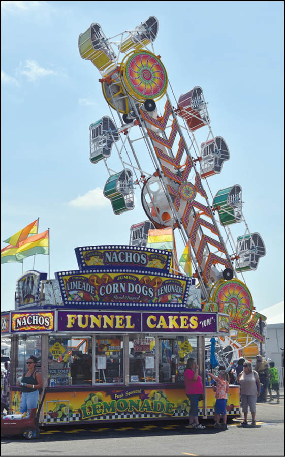 A colorful carnival scene shows a funnel cake and lemonade stand with people standing in front while a large amusement ride with multiple seats is tilted in the background against a clear sky. Description generated by AI