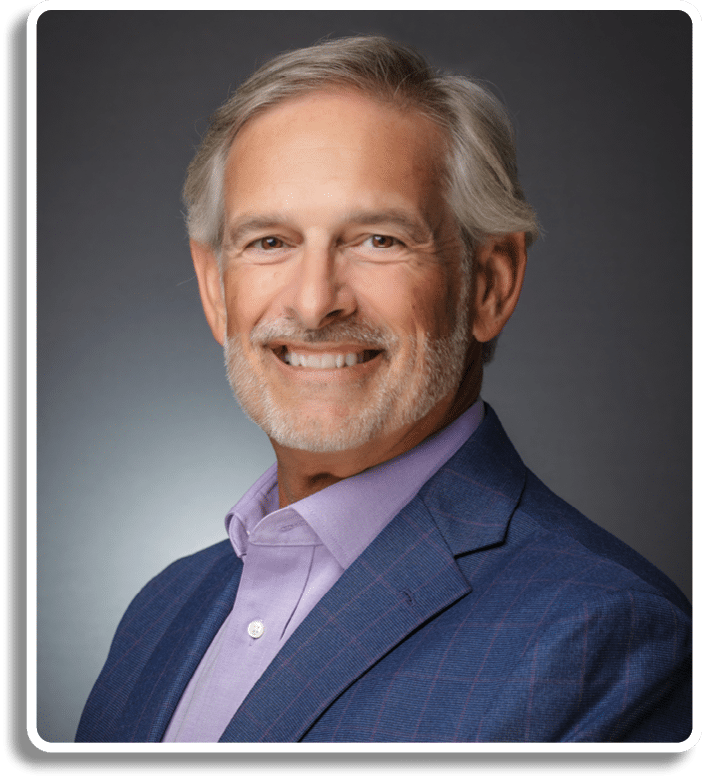 A senior person with gray hair and a beard is smiling while wearing a purple shirt and a blue blazer against a dark gray background. Description generated by AI