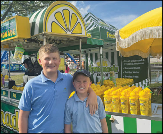 Two children wearing blue shirts stand smiling with one arm around the other in front of a colorful lemonade stand with yellow cups and a yellow umbrella. Description generated by AI