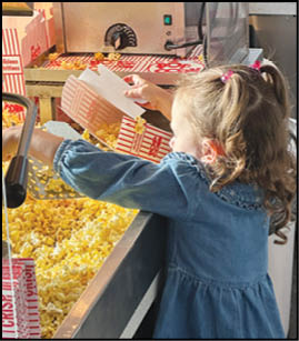 A child with curly hair in a blue dress is scooping popcorn from a large popcorn machine into a striped popcorn box. Description generated by AI