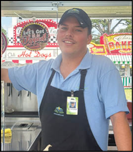 A person wearing a blue polo shirt, black apron, and black cap is standing and smiling inside a food stall with signs advertising hot dogs and baked goods in the background. Description generated by AI
