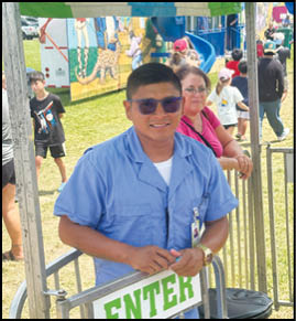 A person wearing sunglasses and a blue uniform stands behind a metal barrier with a sign that says “ENTER" while other people are visible in the background at an outdoor event. Description generated by AI