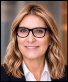 Person with shoulder-length wavy hair wearing black glasses and a dark blazer over a white collared shirt smiling against a blurred background. Description generated by AI