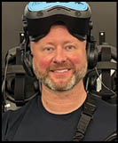 A person wearing a headset and a blue shirt with the word “ZERO" is holding a futuristic black and blue laser tag gun with a digital display while smiling in front of a wall with mounted laser tag equipment. Description generated by AI
