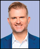 A person with short hair and a trimmed beard is smiling and wearing a blue suit jacket over a white collared shirt against a light blue background. Description generated by AI