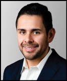 A person with short dark hair and facial hair is smiling and wearing a dark blazer over a white collared shirt against a plain light background. Description generated by AI