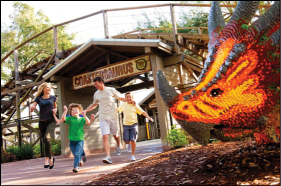 A group of people joyfully walking near a wooden roller coaster named Coastersaurus with a large colorful dinosaur sculpture made of blocks in the foreground. Description generated by AI