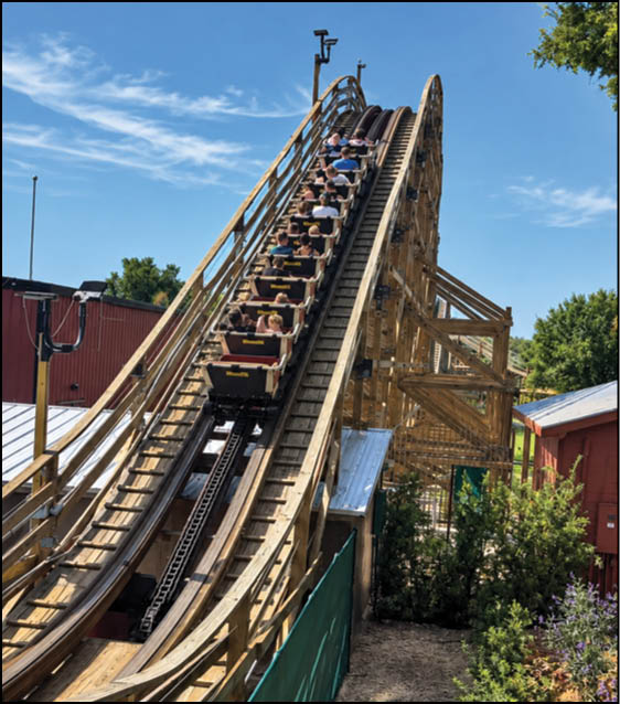 A group of people is seated in a wooden roller coaster car ascending a steep track on a clear, sunny day. Description generated by AI