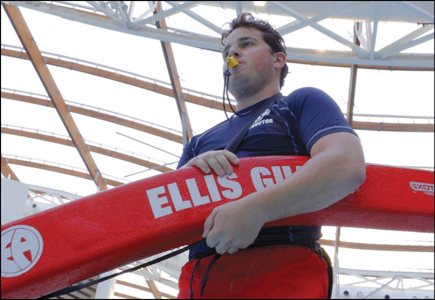 A person wearing a whistle and holding a red rescue float labeled “ELLIS GU" stands indoors under a metal and glass roof. Description generated by AI