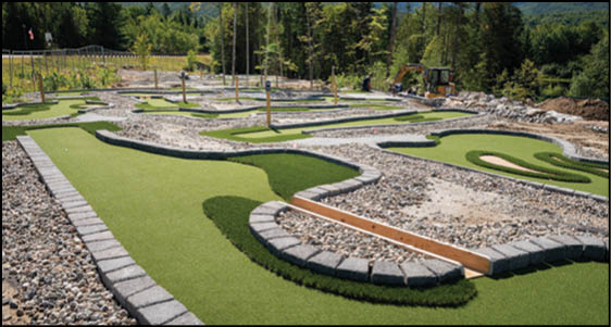 A landscaped outdoor mini golf course with artificial turf, stone borders, sand traps, and trees in the background. Description generated by AI