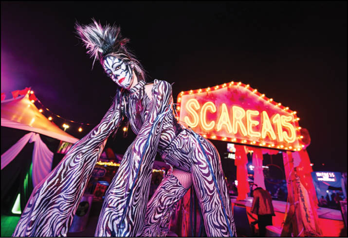 A person in a zebra-striped costume with dramatic hair and face paint poses in front of a brightly lit sign that reads “SCAREA15" at night. Description generated by AI