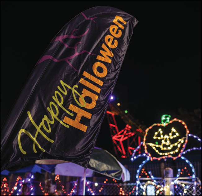 A black flag with the words Happy Halloween in yellow and orange is displayed at night with a background of colorful Halloween-themed lights including a pumpkin face. Description generated by AI