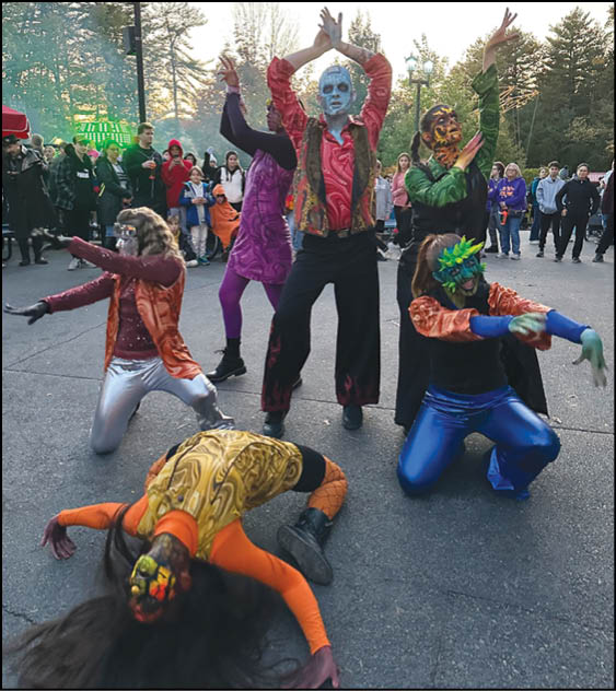 People in colorful costumes and face paint are performing a dramatic dance or theatrical pose outdoors with a crowd watching in the background. Description generated by AI