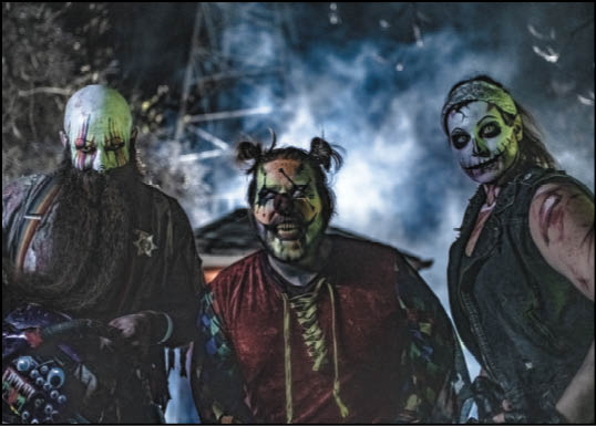 Three people with painted faces and costumes stand outdoors at night with a smoky background, evoking a spooky or Halloween theme. Description generated by AI