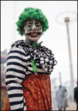 A person dressed as a clown with green curly hair, striped shirt, checkered ruffled collar, and orange polka dot pants stands outdoors with a blurred background of people and structures. Description generated by AI