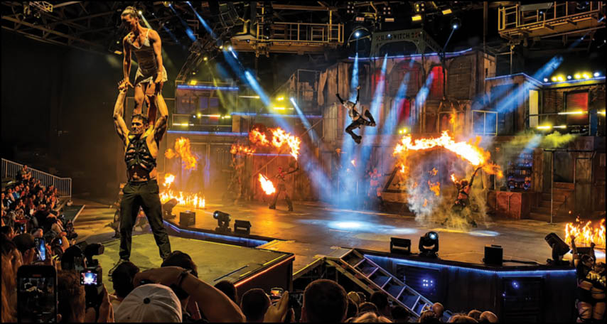 Two adults perform an acrobatic balancing act on stage with dramatic lighting and fire effects while an audience watches and records the show. Description generated by AI