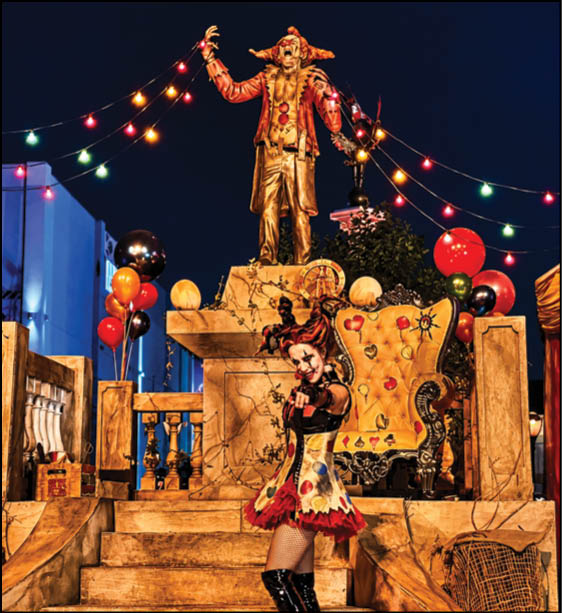 A person dressed in a colorful jester costume poses in front of a decorated stage with a large ornate chair, balloons, string lights, and a golden statue holding a mask. Description generated by AI