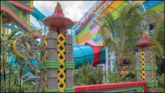 A colorful water park scene with large water slides, tropical plants, and decorative stone pillars with flame sculptures. Description generated by AI