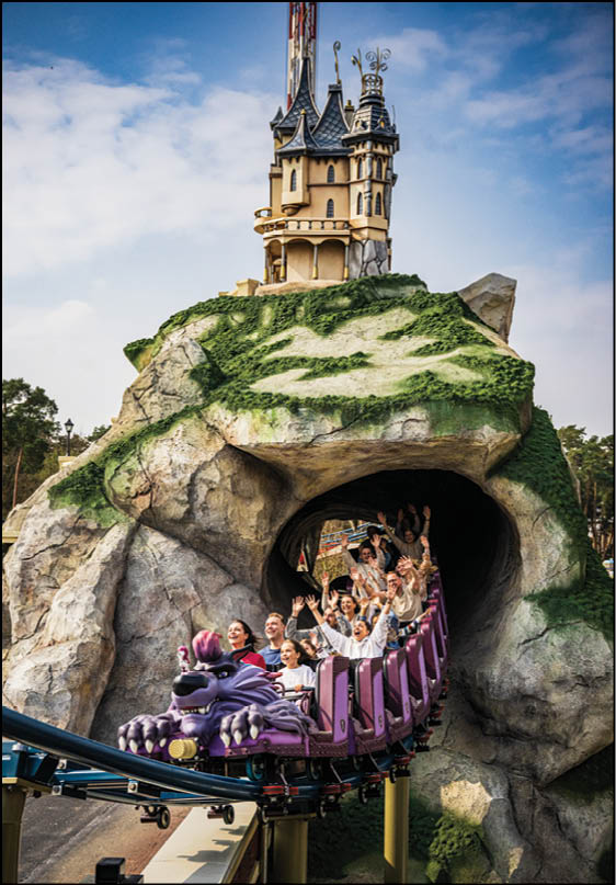 A group of people riding a purple roller coaster train emerging from a tunnel built into a rocky hill with a whimsical castle structure on top under a partly cloudy sky. Description generated by AI