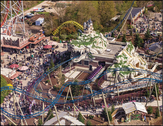 A crowded amusement park features multiple roller coasters weaving through a mountainous castle-themed structure with many people walking and waiting in lines. Description generated by AI