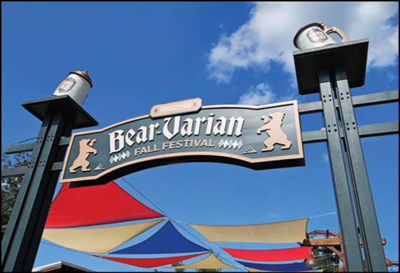 A decorative sign reading “Bear-Varian Fall Festival" with bear silhouettes is mounted on tall posts above colorful red, blue, and beige fabric canopies against a blue sky. Description generated by AI