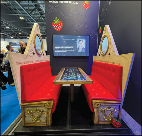 A booth setup with two red cushioned bench seats facing each other across a table with digital screens, decorated with strawberry graphics and a screen displaying a person's photo and text on a dark background. Description generated by AI