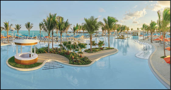 A large outdoor pool area with palm trees, lounge chairs, umbrellas, and a small round cabana surrounded by water under a partly cloudy sky. Description generated by AI
