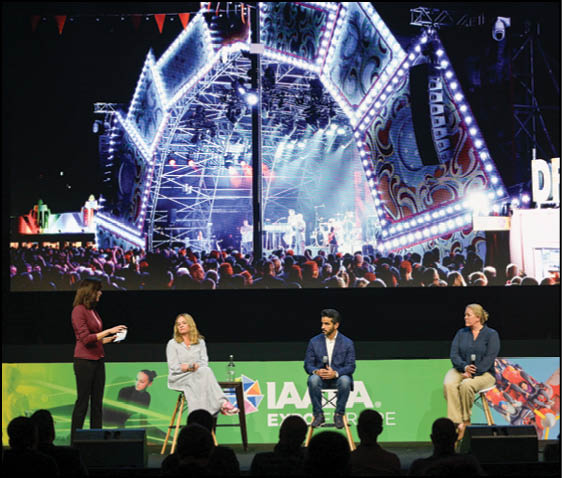 Four adults are seated on stools on a stage with a large screen behind them showing a brightly lit concert scene with a crowd. Description generated by AI