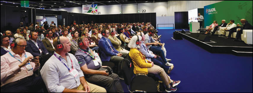 A large group of people wearing headsets is seated attentively in rows facing a stage where several people are seated and one person is speaking at a podium in a conference setting. Description generated by AI